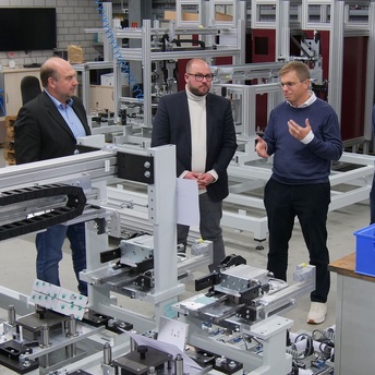 Martin Schmitz, Sebastian Straubel und Alexander Raps stehen vor Teilen einer Maschine und unterhalten sich.