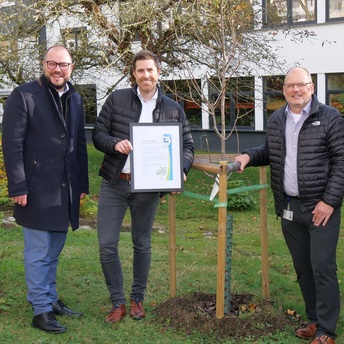Drei Personen stehen vor einem frisch gepflanzten Baum, ein Mann hält eine Urkunde in Händen.