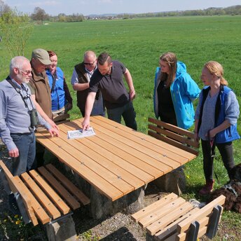 Sieben Personen stehen an einer Sitzgruppe unter freiem Himmel, auf dem Tisch liegt eine Landkarte, im Hintergrund öffnen sich die grünen Wiesen des Tales im Itzgrund.