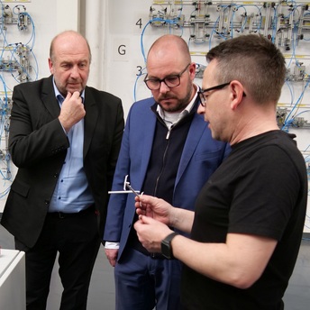 Produktionsleiter Mike Stößel zeigt Martin Schmitz und Landrat Sebastian Straubel ein Kunststoffbauteil. Im Hintergrund ist eine Wand mit vielen elektronischen Bauteilen zu sehen.