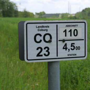 Ein weißes Schild mit der Aufschrift CO23 am Straßenrand. Im Hintergrund sind die Windräder auf den Langen Bergen zu erkennen.