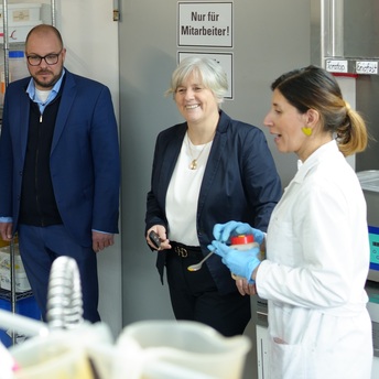 Drei Personen stehen in einem Labor. Rechts eine Frau in einem weißen Laborkittel mit einer Chemikaliendose in der Hand. Sie erklärt, was sie gerade macht.