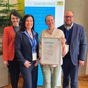 Vanessa Kaiser, Anja Zietz, Kathrin Jouma und Sebastian Straubel stehen vor einem Banner des Freistaats Bayern mit der Urkunde des Wettbewerbes "Innovative Pflege" in der Hand.