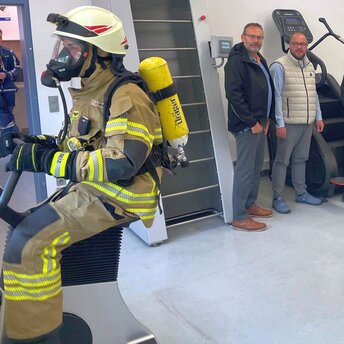 Ein Feuerwehrmann mit Atemschutzausrüstung trainiert auf einem Fahrradergometer. Mehrere Personen in Zivil- und Feuerwehr-Kleidung schauen ihm zu.