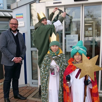 Drei Sternsinger und Landrat Sebastian Straubel stehen vor dem Haupteingang am Landratsamt Coburg. Einer der Sternsinger befestigt den Aufkleber mit dem Segensspruch über der Tür.