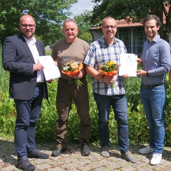 Gruppenbild mit vier Personen vor dem Teich am Landratsamt Coburg. Zwei der Personen halten eine Urkunde für die Bestellung zum Naturschutzwächter in der Hand.