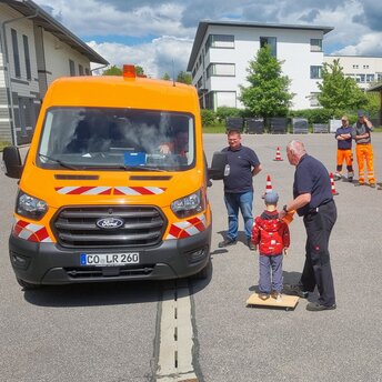 Zwei Männer mit blauen T-Shirt stellen eine Kinderpuppe in den toten Winkel vor einem orangefarbenen Kleinbus.