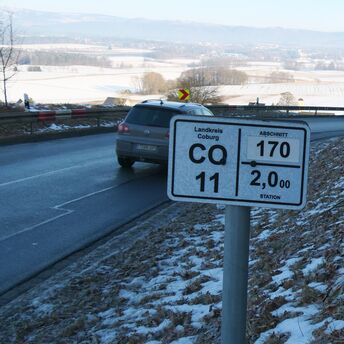 Ein weißes Schild am Rande einer Straße mit der Aufschrift CO 11. Im Hintergrund fährt ein Auto.