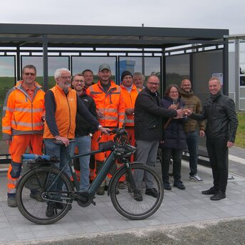 Eine größere Personengruppe, darunter Männer mit orangefarbener Arbeitskleidung, stehen vor einer überdachten Abstellanlage für Fahrräder. Einer der Männer hat ein Fahrrad dabei.