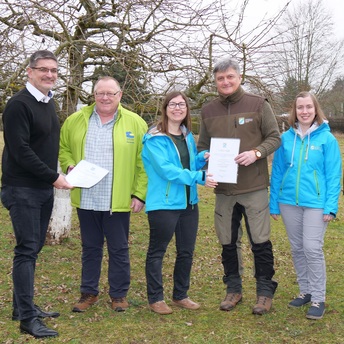 Gruppenbild mit fünf Personen in der freien Natur. Zwei Männer stehen in der Mitte und bekommen ihre Ernennungsurkunden überreicht.