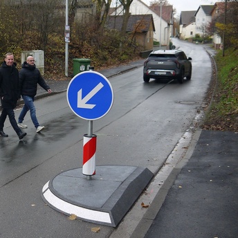 Zwei Personen überqueren eine Straße, im Hintergrund fährt ein Fahrzeug davon. Vorne sind eine Verkehrsinsel am Fahrbahnrand sowie Schild mit einem blauen Hinweispfeil zu sehen.