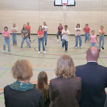 Vier Personen im Vordergrund schauen zu, wie Schülerinnen im Hintergrund einen Tanzt aufführen.