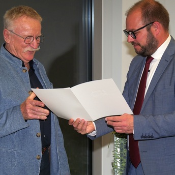 Landrat Sebastian Straubel steht rechts und überreicht an Gerhard Baudler einen Umschlag mit der Kommunalen Dankurkunde