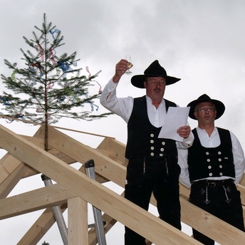 Zimmermann Peter Kraft steht auf dem Dach eines Holzgebäudes. Er hält in der erhobenen Hand ein Glas Sekt und liest von einem Blatt Papier den Richtspruch